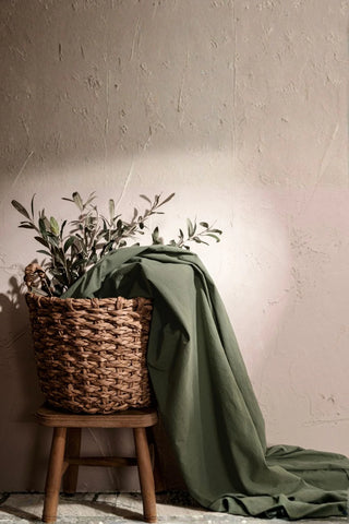 Dark green stonewashed cotton curtain fabric draped over a basket with olive branches. Handwoven in Denizli, OEKO-TEX. Vivamaison.