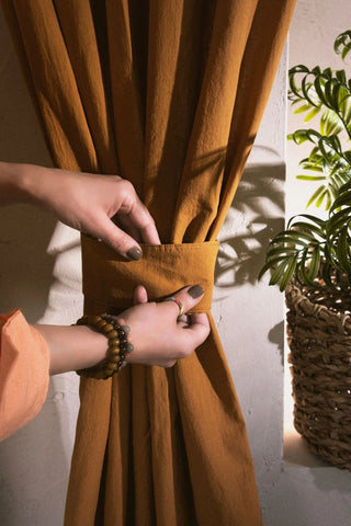 Hands fastening the tieback band of a terracotta stonewashed cotton curtain. Handwoven in Denizli, OEKO-TEX. Vivamaison.