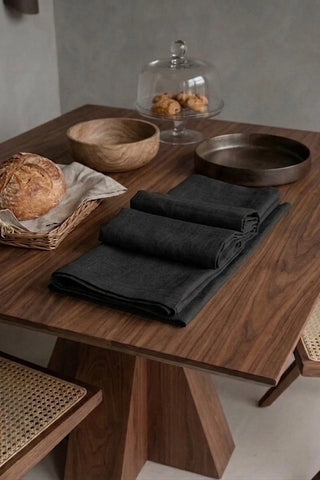 Black plain linen cotton blend table runner folded on a walnut dining table beside a bread basket. 16 x 51 in, handmade in Denizli, OEKO-TEX. VivaMaison.