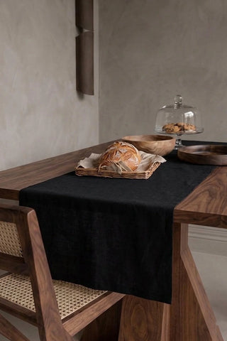 Black plain linen cotton blend table runner draped over a walnut dining table with a bread basket and wooden bowls. 16 x 51 in, handmade in Denizli, OEKO-TEX. VivaMaison.