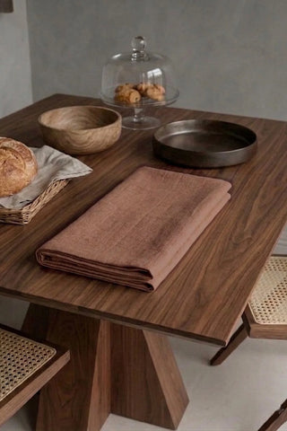 Brown plain linen cotton blend table runner folded on a walnut dining table beside a bread basket. 16 x 51 in, handmade in Denizli, OEKO-TEX. VivaMaison.