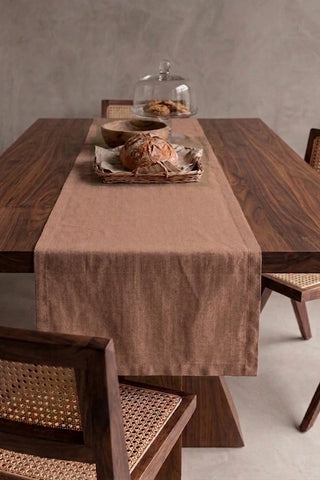 Brown plain linen cotton blend table runner draped over a walnut dining table with a bread basket and wooden bowls. 16 x 51 in, handmade in Denizli, OEKO-TEX. VivaMaison.