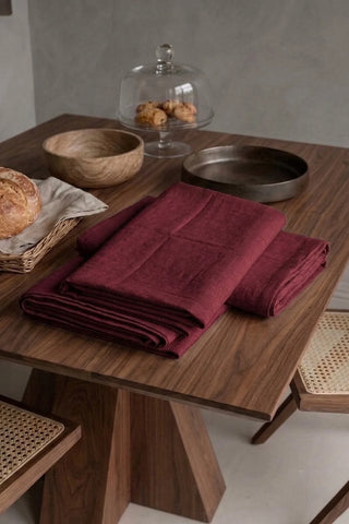 Burgundy plain linen cotton blend table runner folded on a walnut dining table beside a bread basket. 16 x 51 in, handmade in Denizli, OEKO-TEX. VivaMaison.