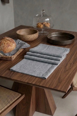 Gray plain linen cotton blend table runner folded on a walnut dining table beside a bread basket. 16 x 51 in, handmade in Denizli, OEKO-TEX. VivaMaison.