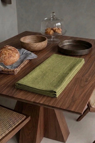 Olive Green plain linen cotton blend table runner folded on a walnut dining table beside a bread basket. 16 x 51 in, handmade in Denizli, OEKO-TEX. VivaMaison.