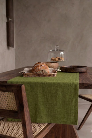 Olive Green plain linen cotton blend table runner draped over a walnut dining table with a bread basket and wooden bowls. 16 x 51 in, handmade in Denizli, OEKO-TEX. VivaMaison.