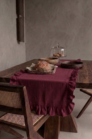 Burgundy ruffled linen cotton blend table runner draped over a walnut dining table with a bread basket and wooden bowls. 16 x 51 in, handmade in Denizli, OEKO-TEX. VivaMaison.