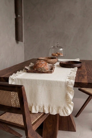 Natural ruffled linen cotton blend table runner draped over a walnut dining table with a bread basket and wooden bowls. 16 x 51 in, handmade in Denizli, OEKO-TEX. VivaMaison.