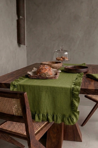 Olive Green ruffled linen cotton blend table runner draped over a walnut dining table with a bread basket and wooden bowls. 16 x 51 in, handmade in Denizli, OEKO-TEX. VivaMaison.
