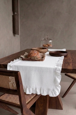 White ruffled linen cotton blend table runner draped over a walnut dining table with a bread basket and wooden bowls. 16 x 51 in, handmade in Denizli, OEKO-TEX. VivaMaison.
