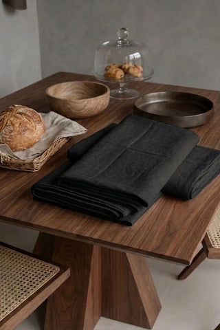 Black plain linen cotton blend tablecloth folded on a walnut dining table beside a bread basket. 9 sizes, handmade in Denizli, OEKO-TEX. VivaMaison.