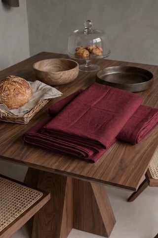 Burgundy plain linen cotton blend tablecloth folded on a walnut dining table beside a bread basket. 9 sizes, handmade in Denizli, OEKO-TEX. VivaMaison.