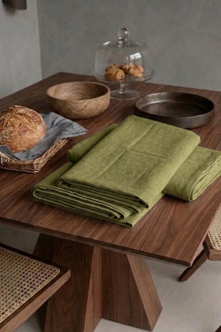 Olive Green plain linen cotton blend tablecloth folded on a walnut dining table beside a bread basket. 9 sizes, handmade in Denizli, OEKO-TEX. VivaMaison.