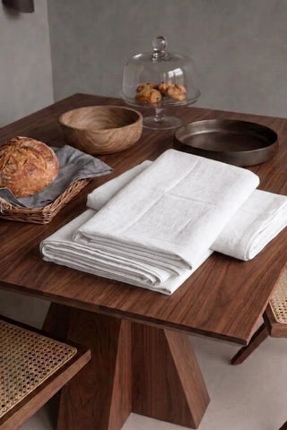 White plain linen cotton blend tablecloth folded on a walnut dining table beside a bread basket. 9 sizes, handmade in Denizli, OEKO-TEX. VivaMaison.