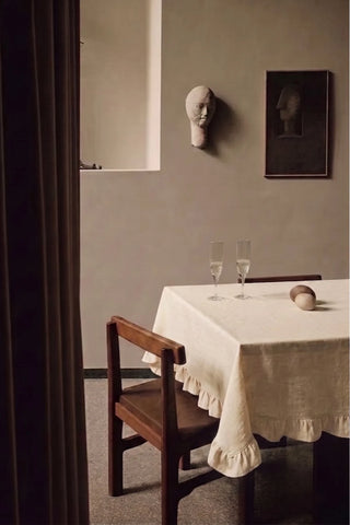 Natural ruffled linen cotton blend tablecloth on a dining table with champagne flutes, seen through a doorway. 9 sizes, handmade in Denizli, OEKO-TEX. VivaMaison.