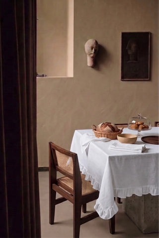White ruffled linen cotton blend tablecloth on a dining table with a bread basket, seen through a doorway. 9 sizes, handmade in Denizli, OEKO-TEX. VivaMaison.